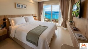 A hotel room with a large bed, a TV, a desk with a chair, and a view of the sea.