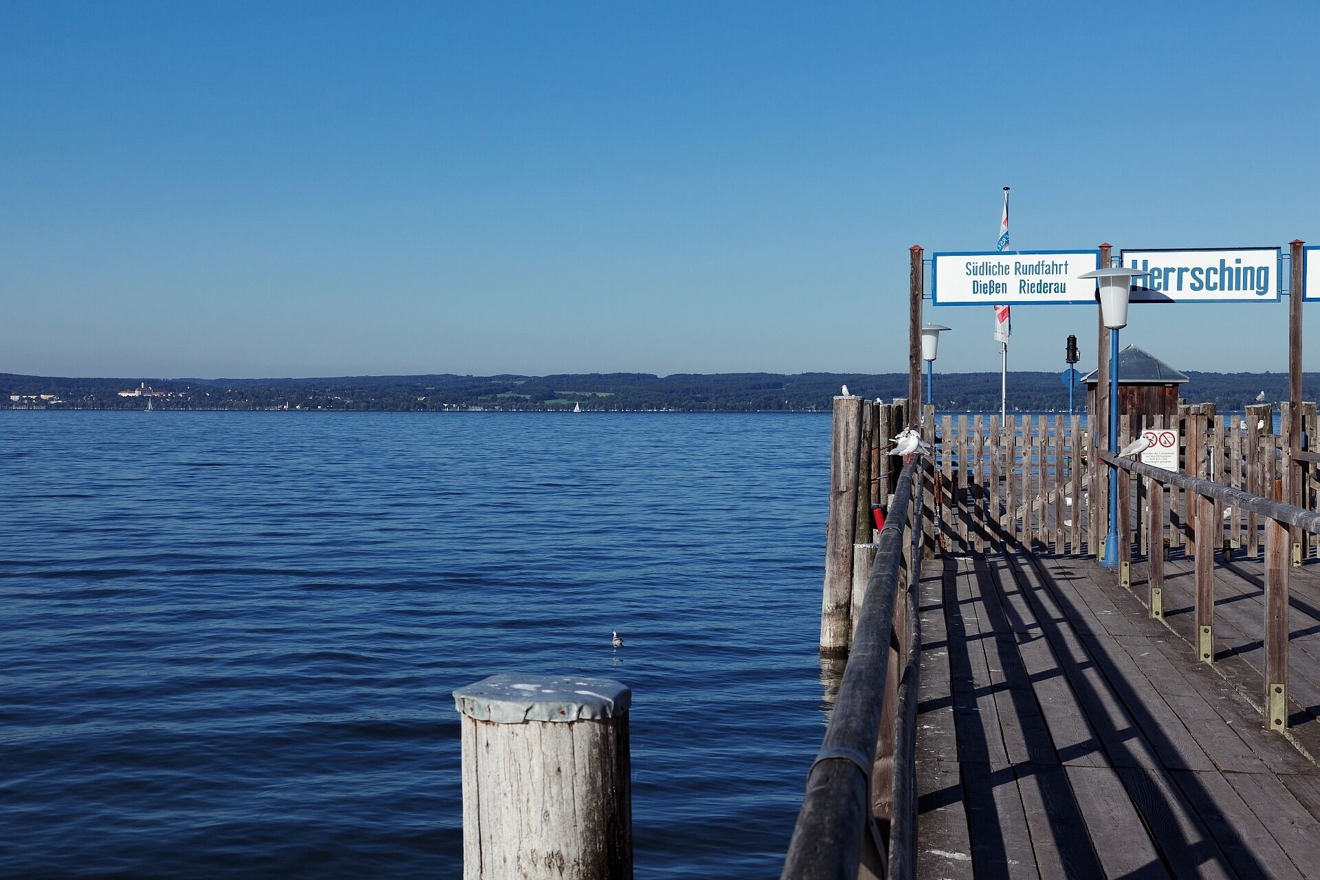 En träbrygga som sträcker sig ut i vattnet, med en klar himmel ovanför. Det finns skyltar som visar vägen till ”Südliche Rundfahrt” och ”Hersching”.