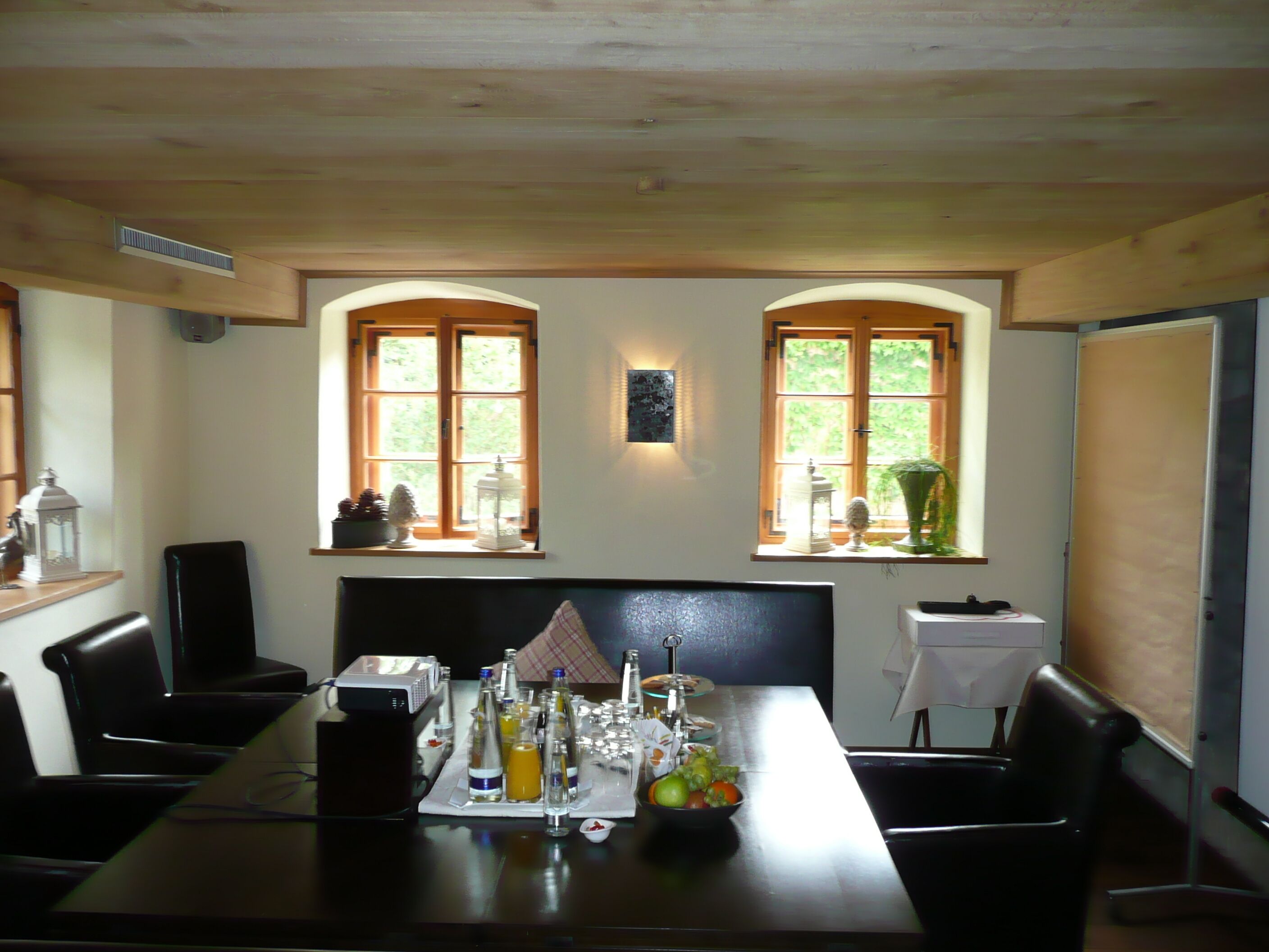 A dining area with a long table, two windows with arched tops, and a view of greenery outside.