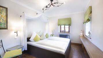 A bedroom with a bed, a chair, a table, a window with curtains, a radiator, and a chandelier.