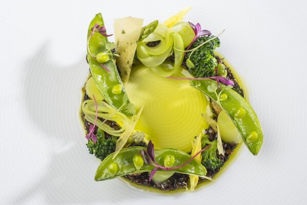 Un plato blanco con un elemento amarillo en el centro, rodeado de verduras verdes y toques morados.