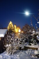 Paisagem coberta de neve com uma igreja iluminada, uma árvore nevada e um muro de pedra.