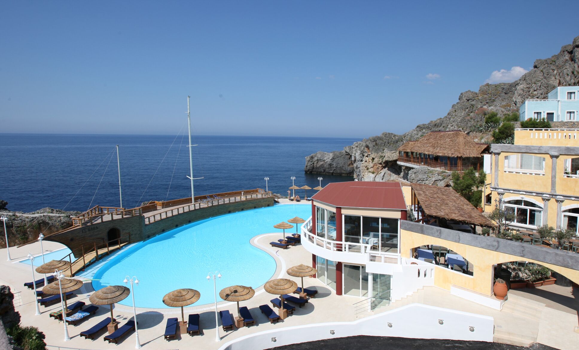 Une piscine de complexe hôtelier avec des chaises longues et des parasols, un bâtiment avec un toit rouge et une vue sur la mer.