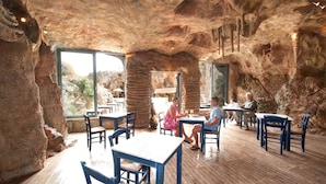A restaurant with a cave-like interior, featuring natural rock formations and large windows. Tables and chairs are arranged for dining.