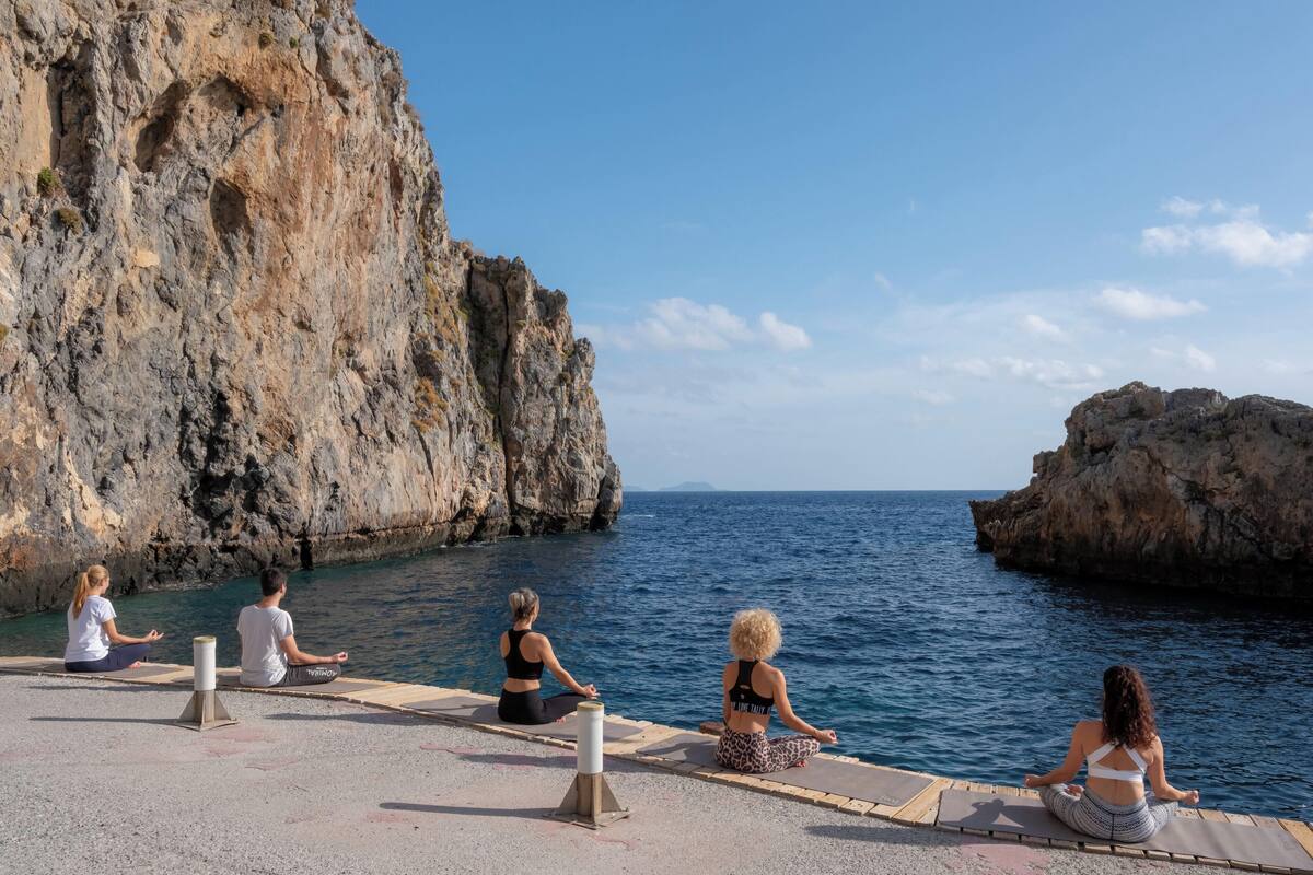 Quatre personnes méditent sur une corniche en béton, au bord d’une côte rocheuse, avec une eau bleue et limpide.