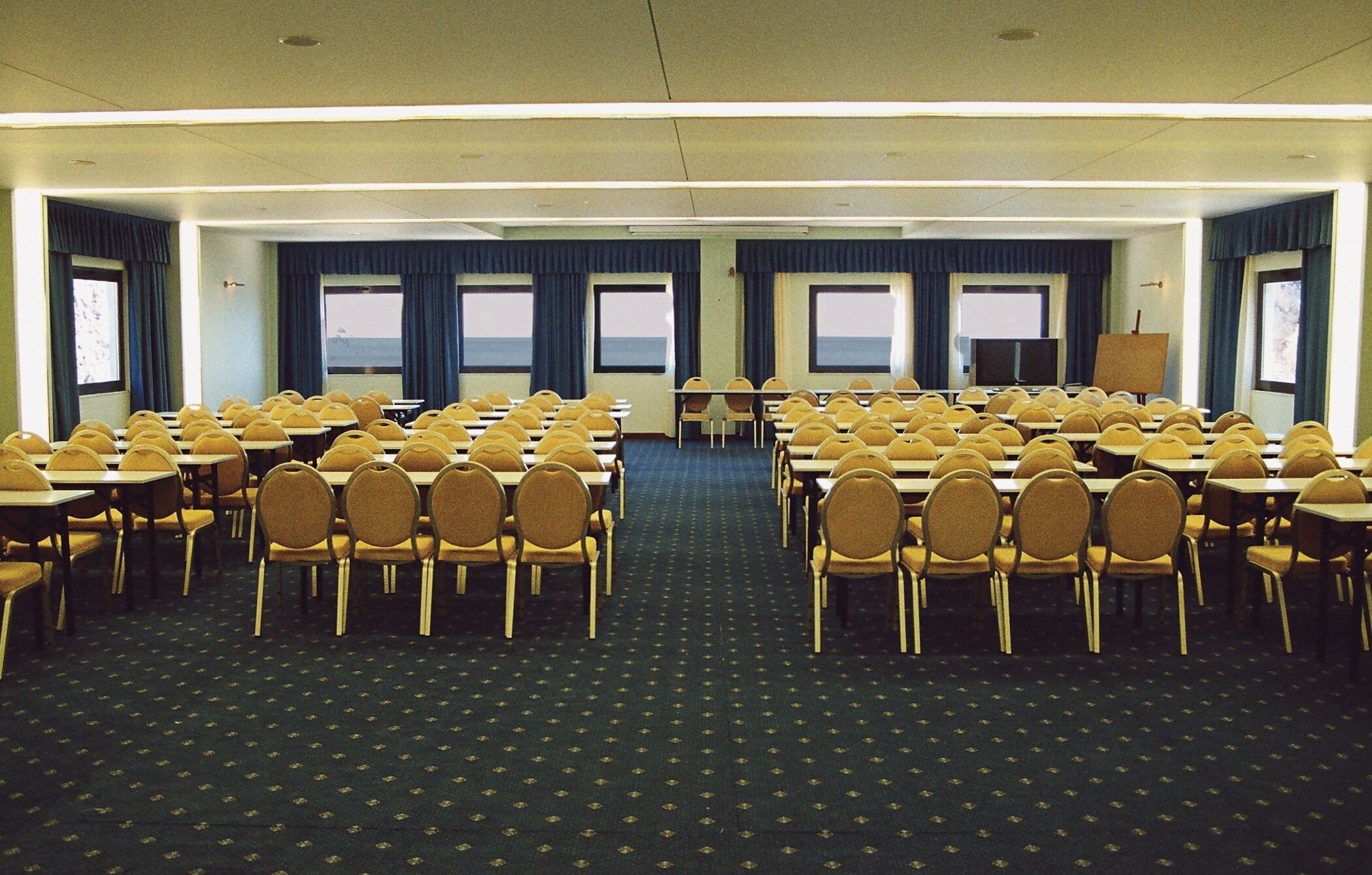 Un salón de conferencias con filas de sillas y mesas, ventanas amplias con cortinas, y suelo alfombrado.