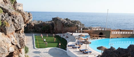 Un área de piscina con vista al mar, una cancha de voleibol y un muelle de madera con sillones de descanso.