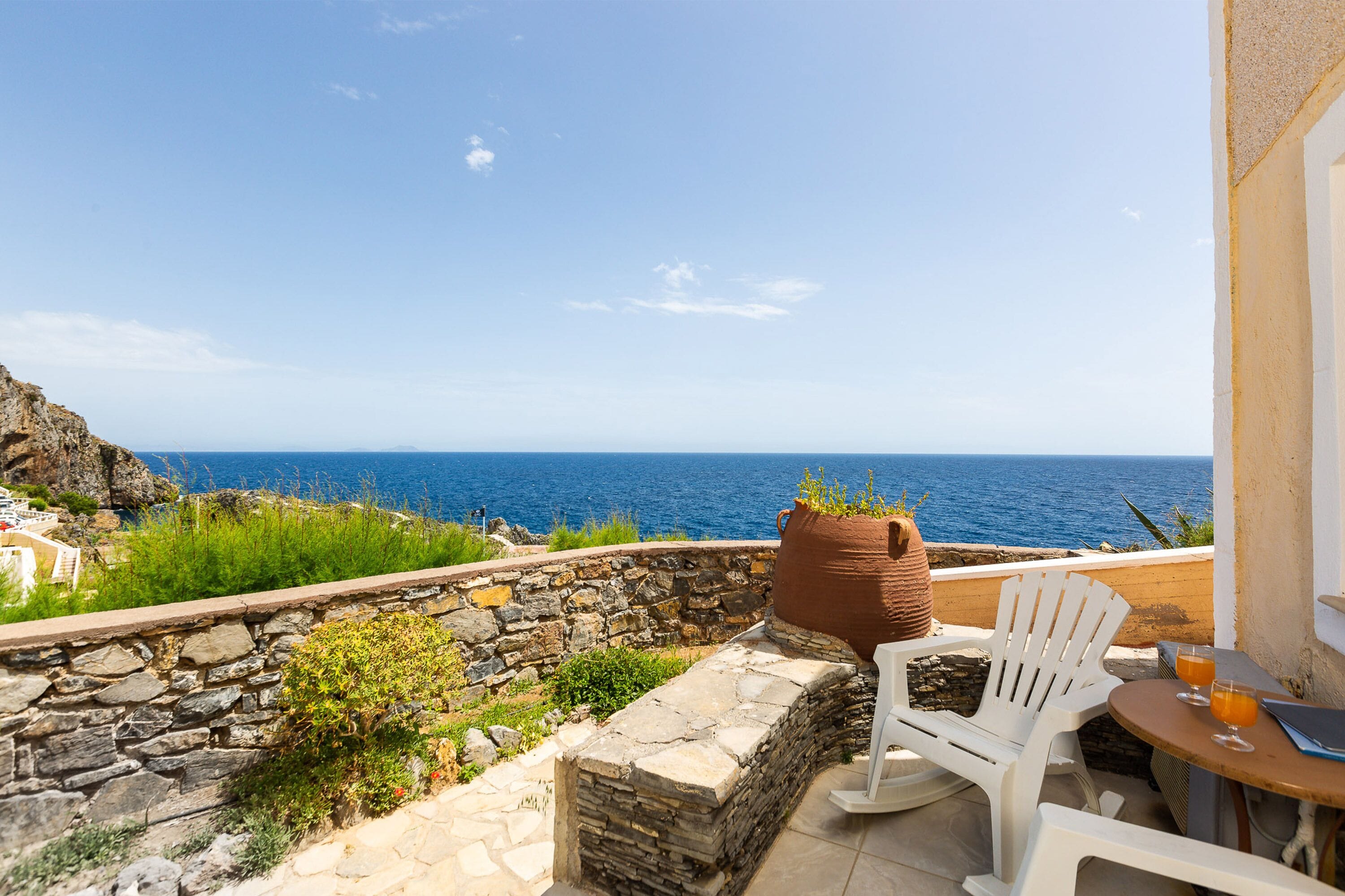 A terrace with a stone wall, white chairs, a table with drinks, and a view of the sea.