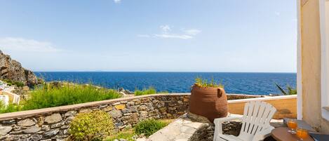 A terrace with a stone wall, white chairs, a table with drinks, and a view of the sea.