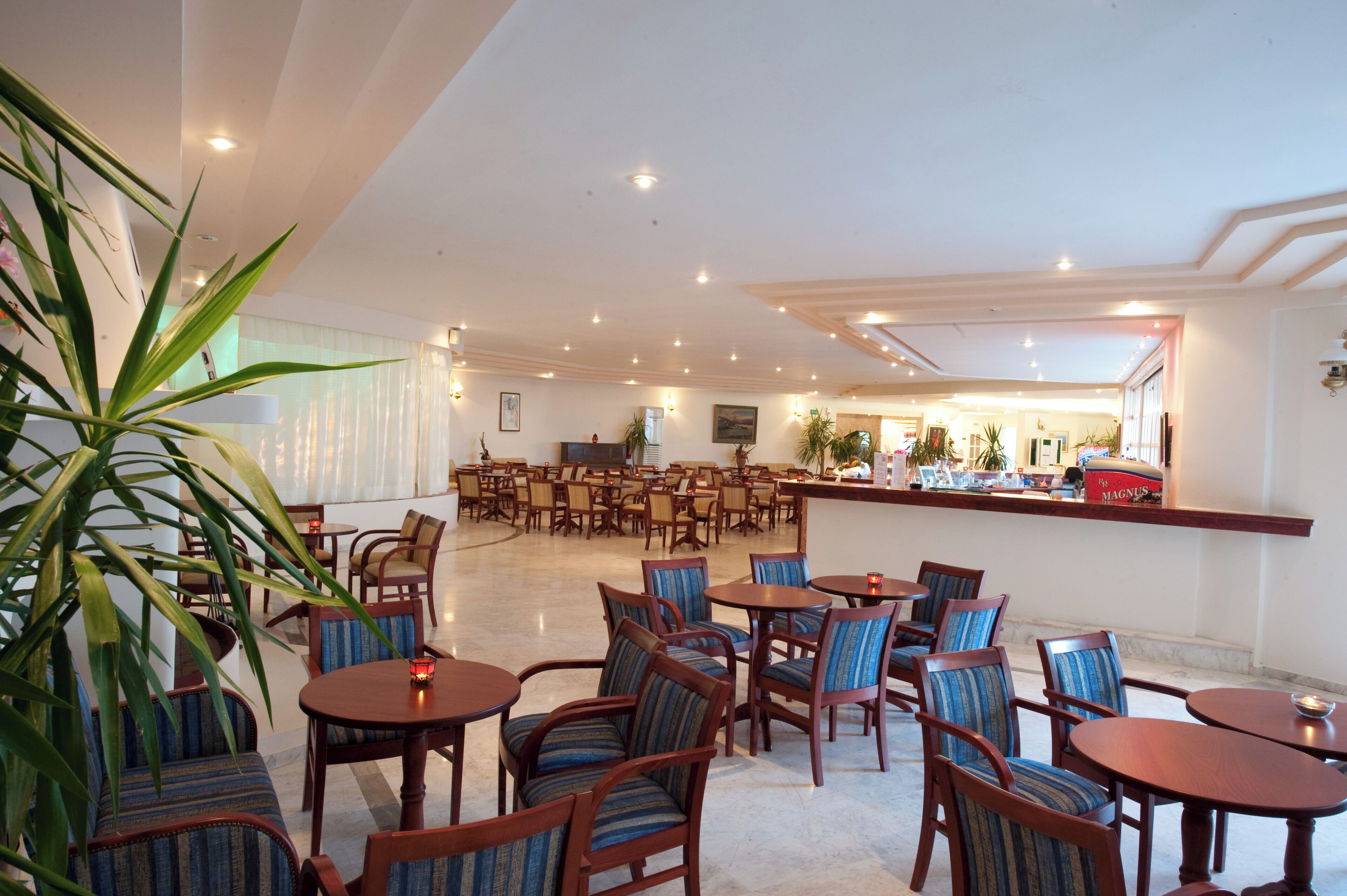 A spacious hotel lobby with multiple seating areas, including round tables and chairs, and a reception desk with a bar.