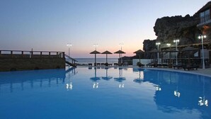 A swimming pool with lounge chairs and umbrellas, a stone wall, and a view of the ocean at dusk.