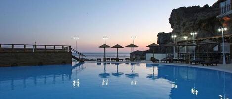 A swimming pool with lounge chairs and umbrellas, a stone wall, and a view of the ocean at dusk.