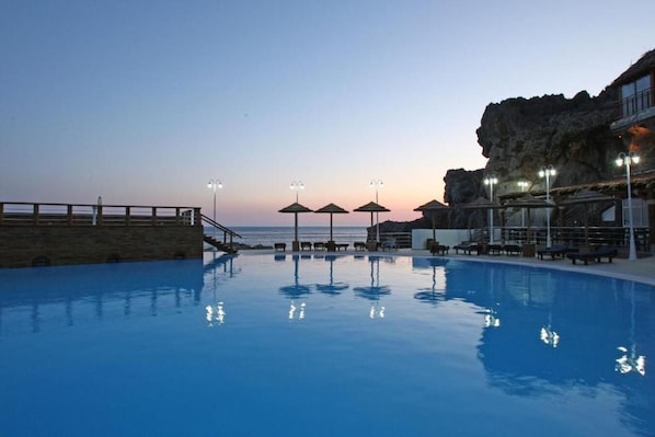 Une piscine avec des chaises longues et des parasols, un mur de pierre et une vue sur l’océan au crépuscule.