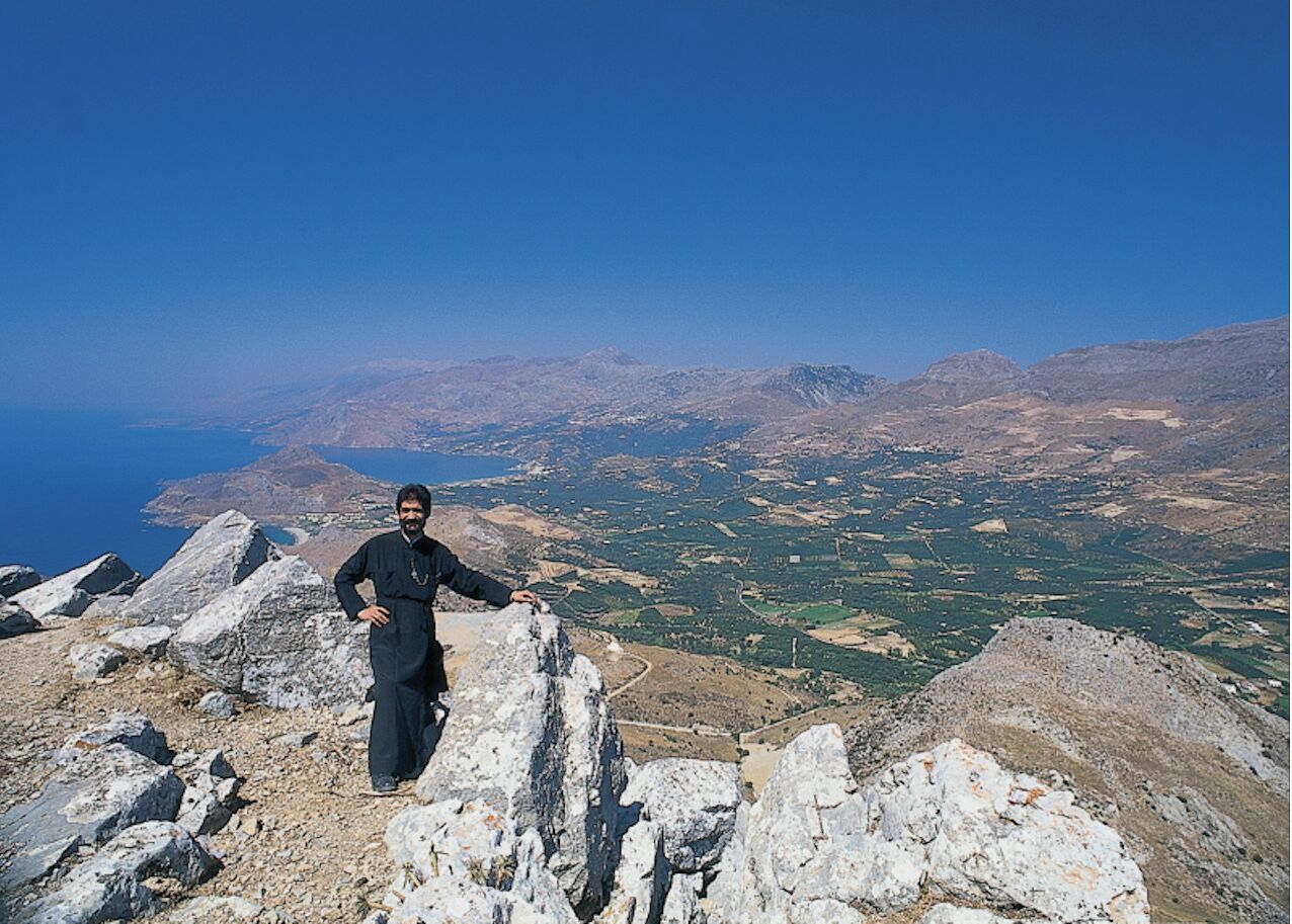 Una persona de pie sobre la cima rocosa de una montaña, con vistas a un valle y montañas lejanas.