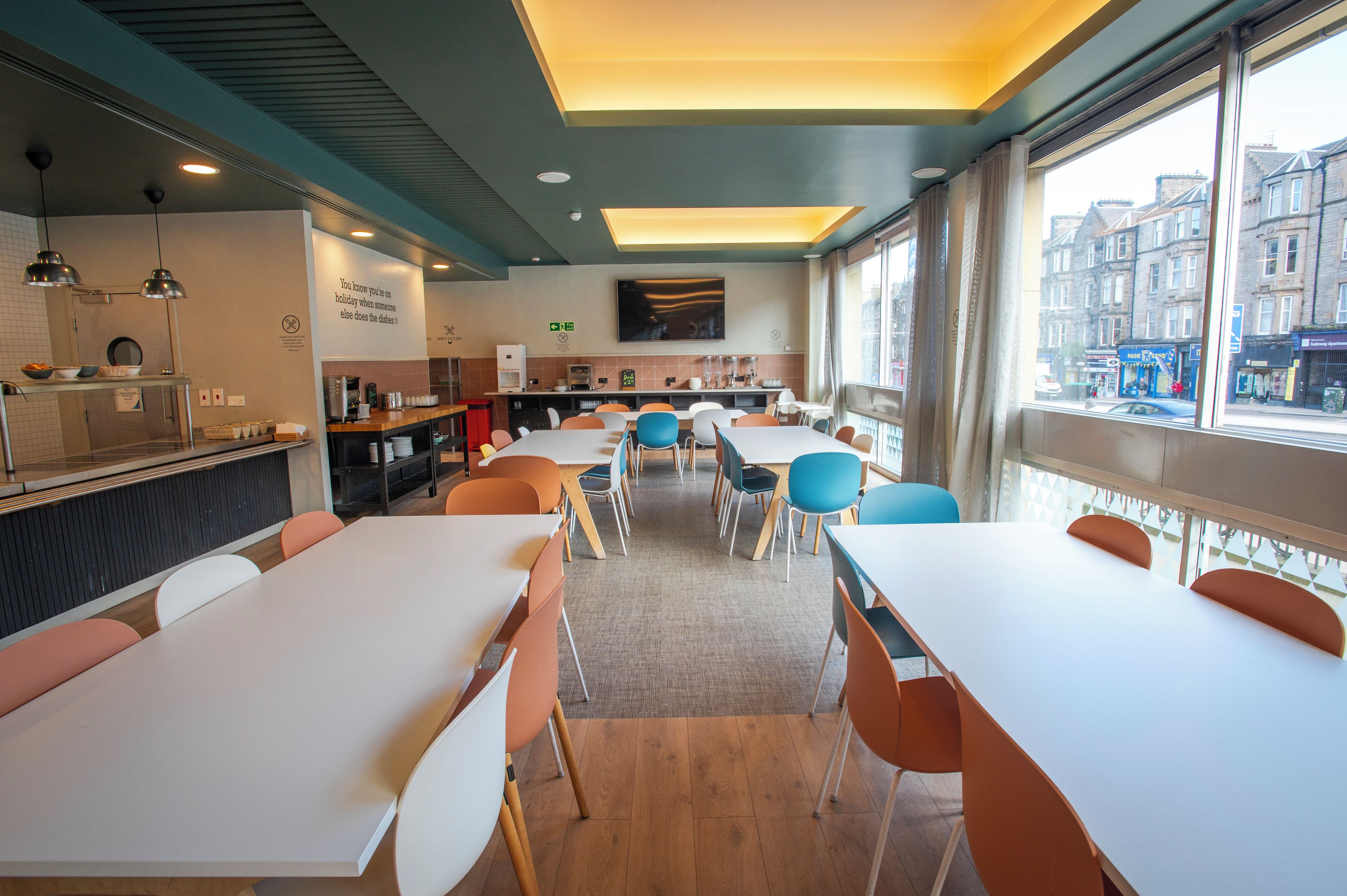 A modern dining area with white tables and orange chairs, a kitchen in the background, and a city view through large windows.