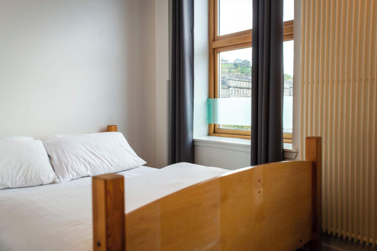 A bedroom with a wooden bed, a window with curtains, and a view of buildings.