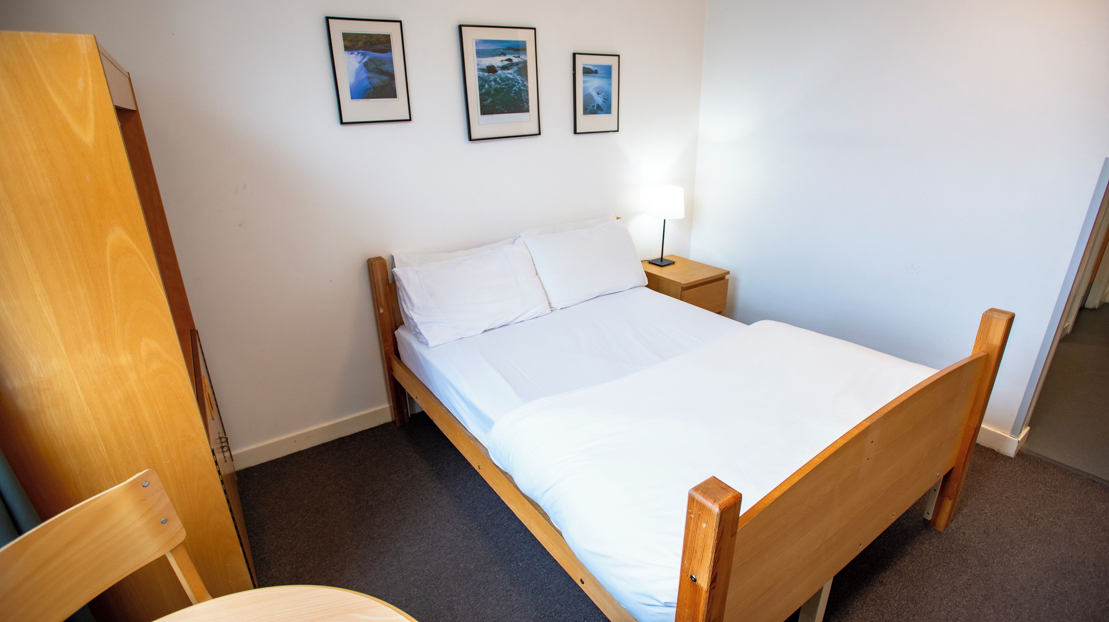 A bedroom with a wooden bed, white bedding, a nightstand with a lamp, and three framed pictures on the wall.