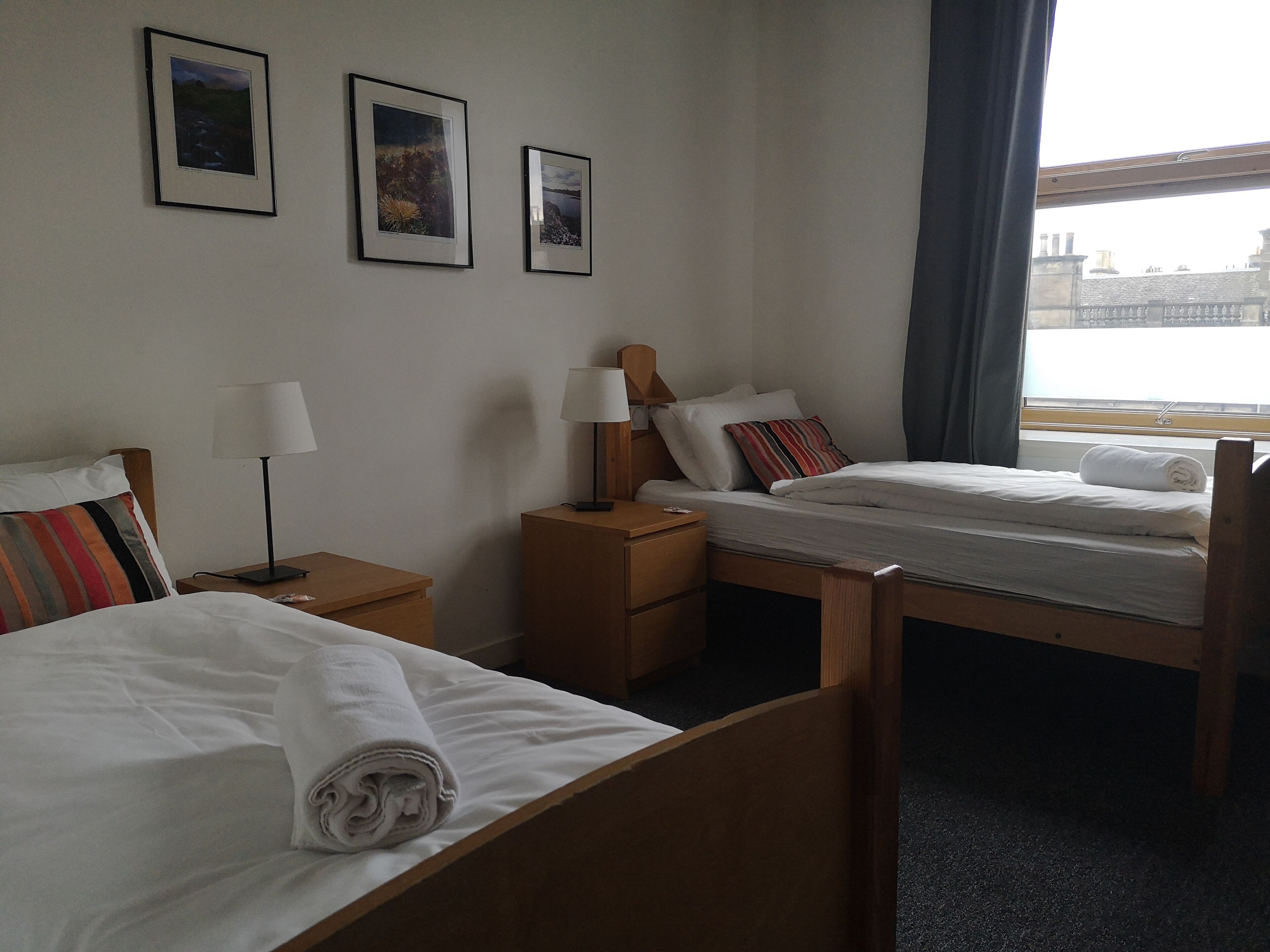 Een hotelkamer met twee bedden, een nachtkastje met een lamp, een bijzettafeltje en een raam met uitzicht op gebouwen.