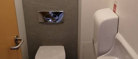 A small, compact bathroom with a toilet, urinal, and a paper towel dispenser.