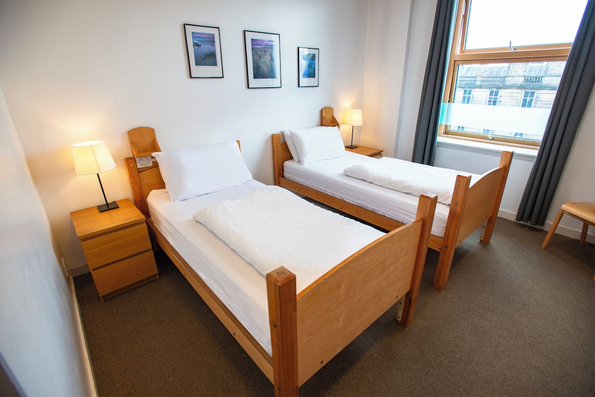 A hotel room with two wooden beds, a nightstand with a lamp, and a window with curtains.