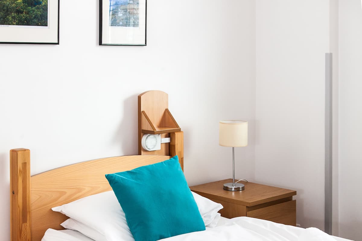 A neatly made bed with white linens and a teal pillow, a wooden headboard, a bedside table with a lamp, and framed pictures on the wall.