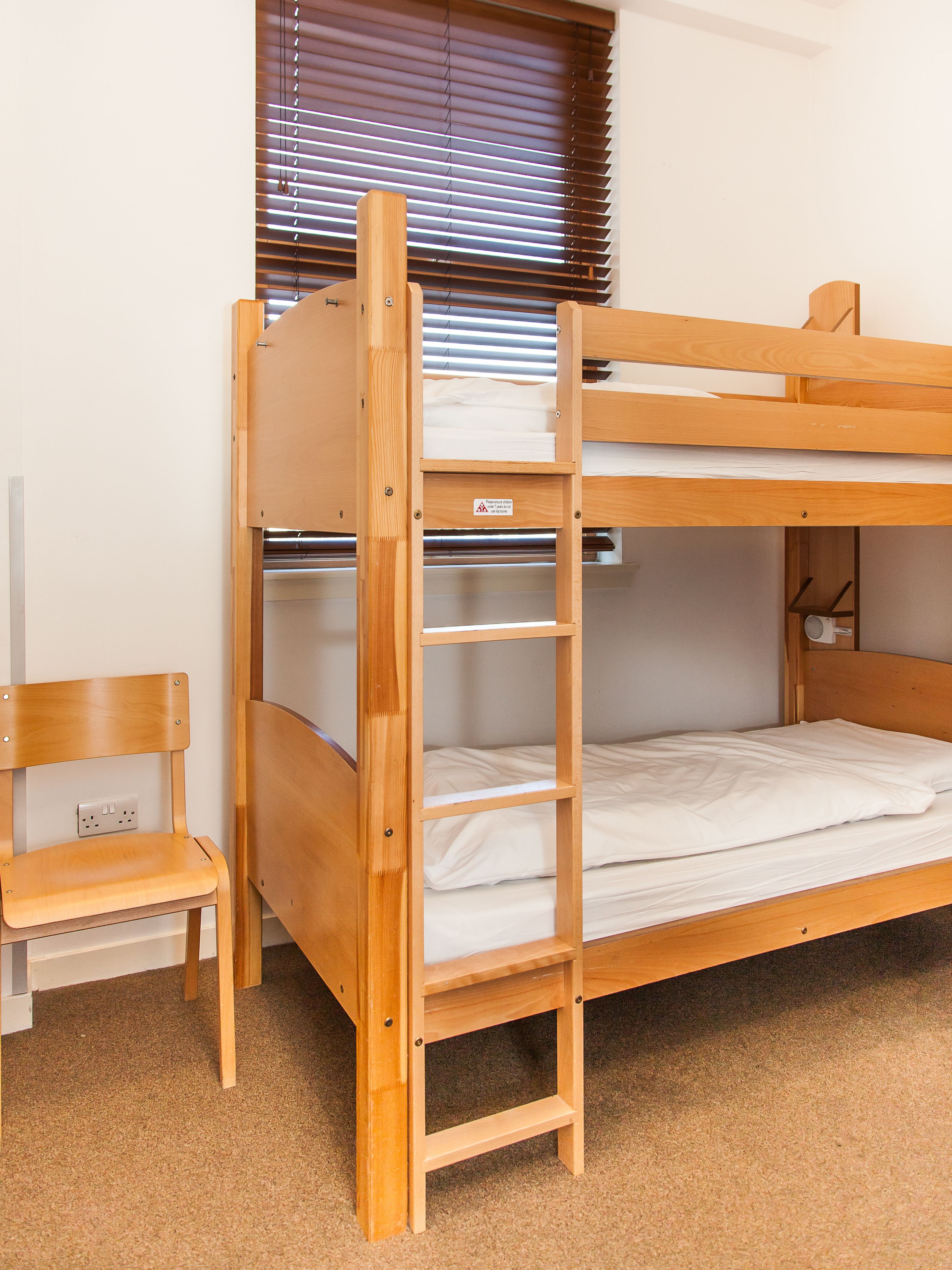 A bunk bed with a wooden ladder, a small wooden chair, and a window with blinds.
