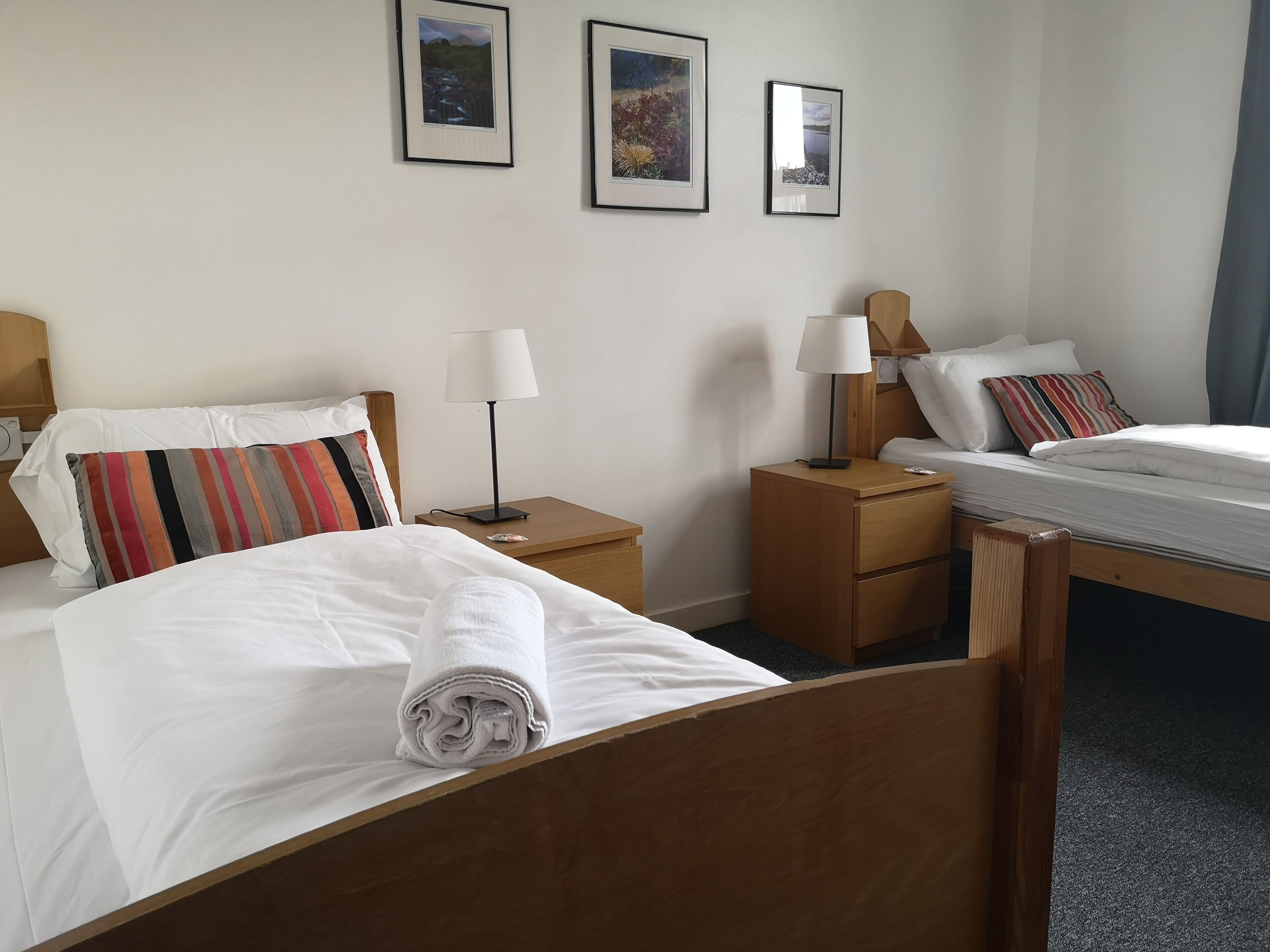 A hotel room with two beds, a wooden headboard, a nightstand with a lamp, and a small table.