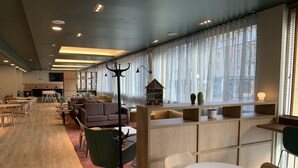 Un hall d’hôtel moderne avec un plancher en bois, des coins salon, un comptoir d’accueil et de grandes fenêtres ornées de rideaux légers.