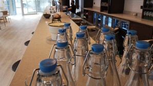 Un espace bar avec plusieurs bouteilles en verre transparent, dotées de bouchons bleus, un bol blanc et une personne à l’arrière-plan.