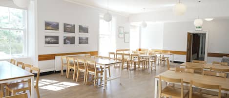 Een ruime kamer met meerdere houten tafels en stoelen, grote ramen en wanddecoraties.