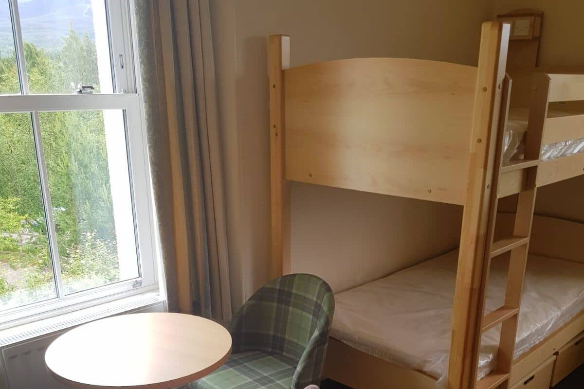 A bunk bed with a wooden ladder, two plaid chairs, and a small round table in a room with a window.