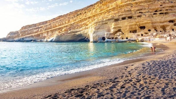 Uma praia com águas azuis cristalinas, areal arenoso e falésia rochosa com grutas.