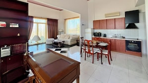 Uma sala de estar ampla com sofá, mesa de centro e área de jantar com cadeiras e mesa. Uma cozinha com armários e eletrodomésticos é visível ao fundo.