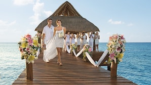 A wooden pier leading to a thatched-roof structure over water, with floral arrangements and guests. - Secrets Aura Cozumel - Adults Only - All Inclusive (Cozumel)