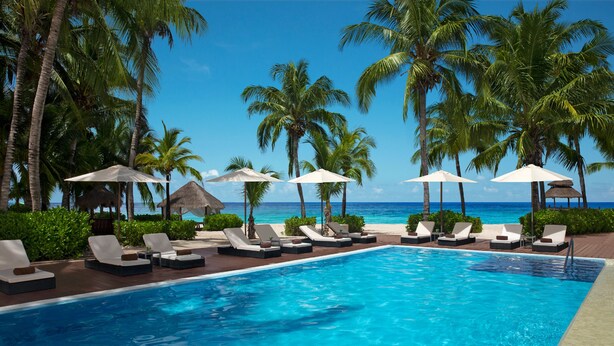 A poolside area with lounge chairs and umbrellas, surrounded by palm trees and a clear view of the ocean. - Secrets Aura Cozumel - Adults Only - All Inclusive (Cozumel)