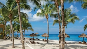 A beach with palm trees, lounge chairs, and thatched umbrellas. - Secrets Aura Cozumel - Adults Only - All Inclusive (Cozumel)