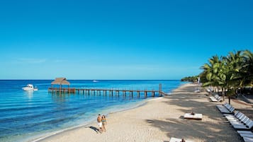 Una playa con muelle de madera, sillas de descanso y una cabaña de techo de paja.