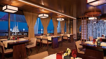 A restaurant with multiple tables, wooden flooring, and large windows offering a view of the cityscape.