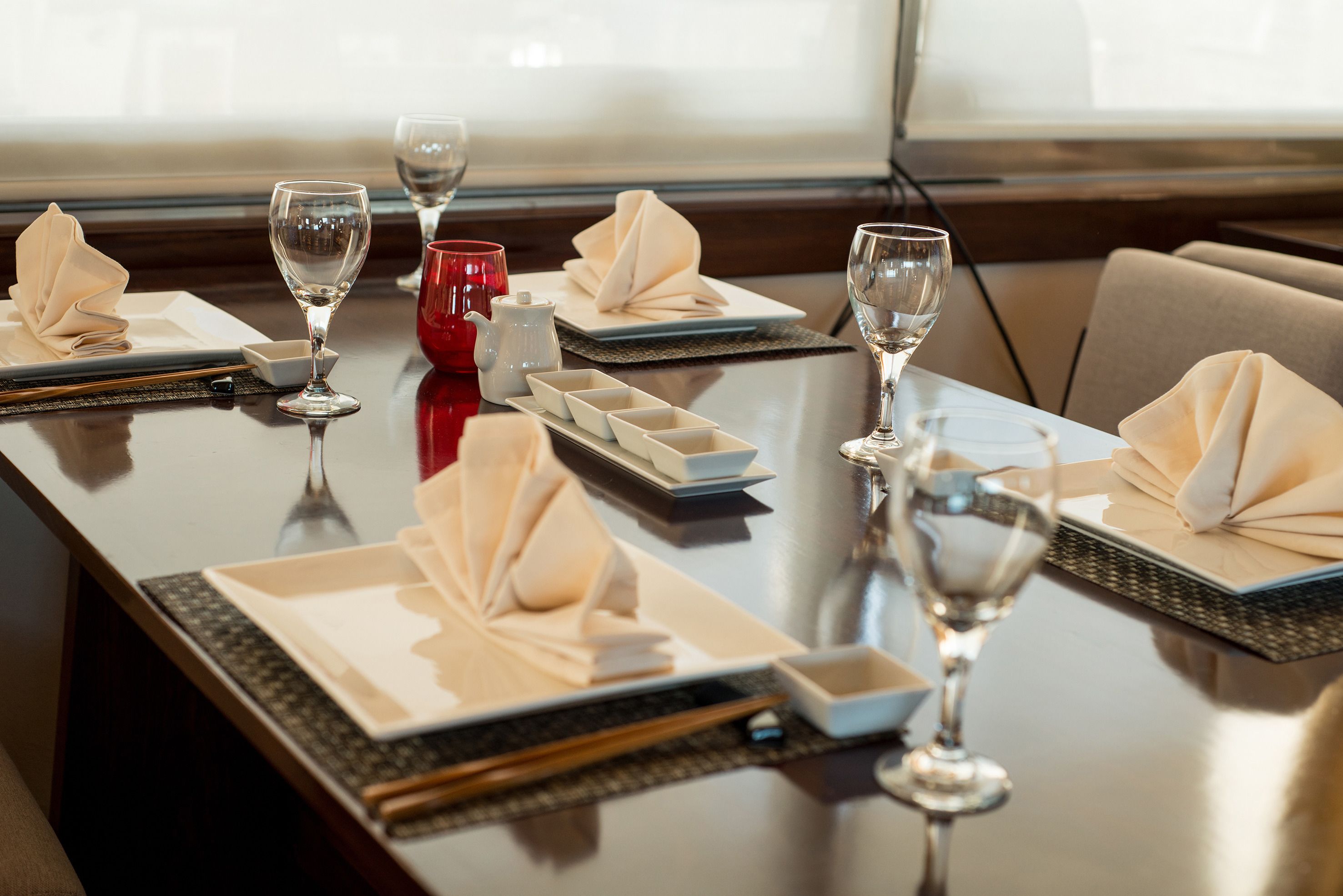 Mesa de comedor dispuesta con platos, servilletas, vasos y una taza roja, lo que sugiere un ambiente formal para comer.
