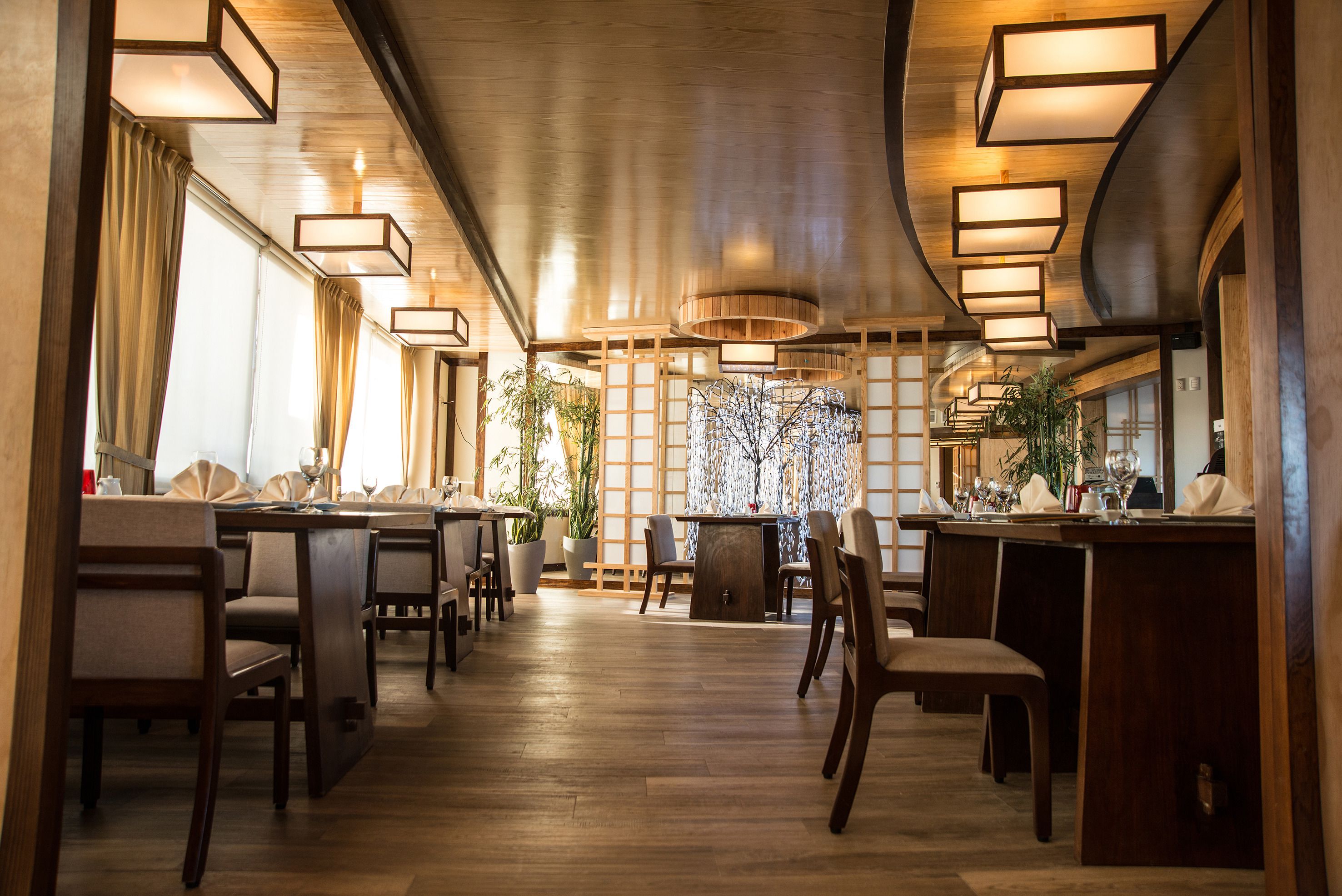 A restaurant with wooden floors, tables, and chairs. There are hanging lights and a plant in the center.