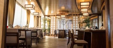 A restaurant with wooden floors, tables, and chairs. There are hanging lights and a plant in the center.