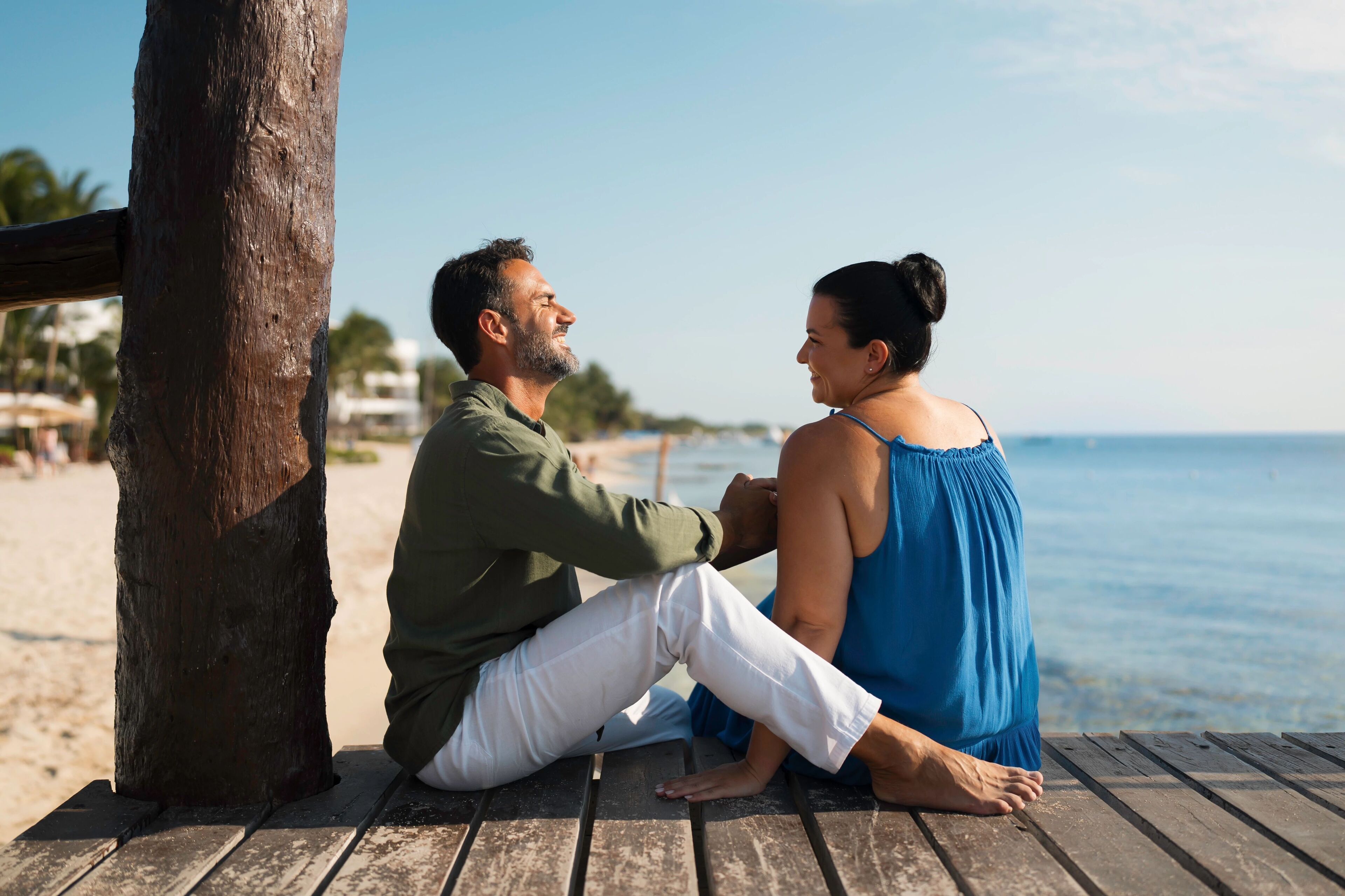A couple sitting on a wooden bench by the beach, with a wooden post and a clear view of the ocean.