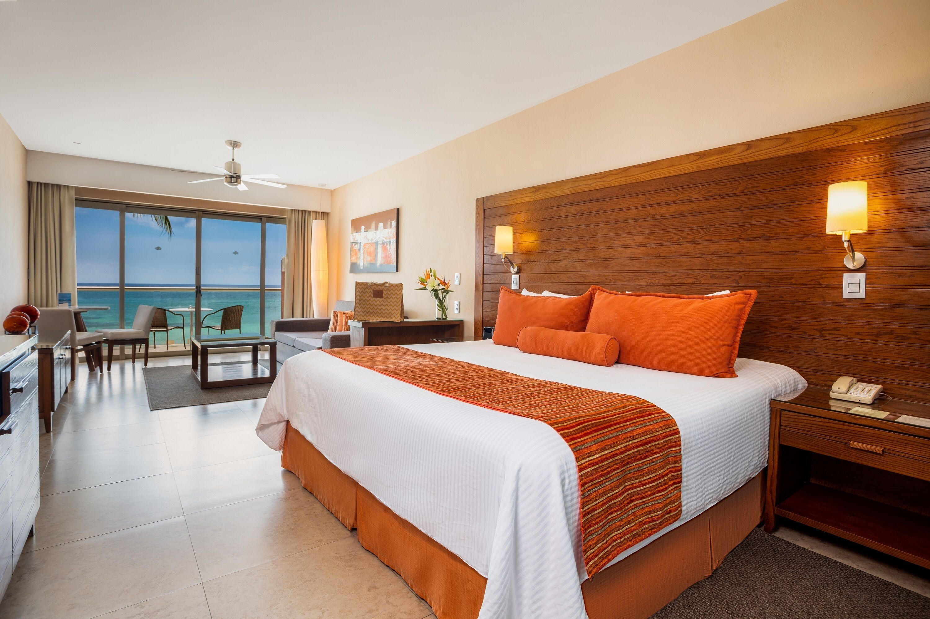 A hotel room with a large bed, a desk, a chair, a small table, and a view of the ocean.