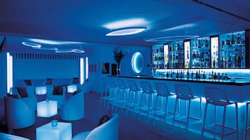 A modern bar area with a blue-lit ambiance, white bar stools, and a variety of bottles on shelves.
