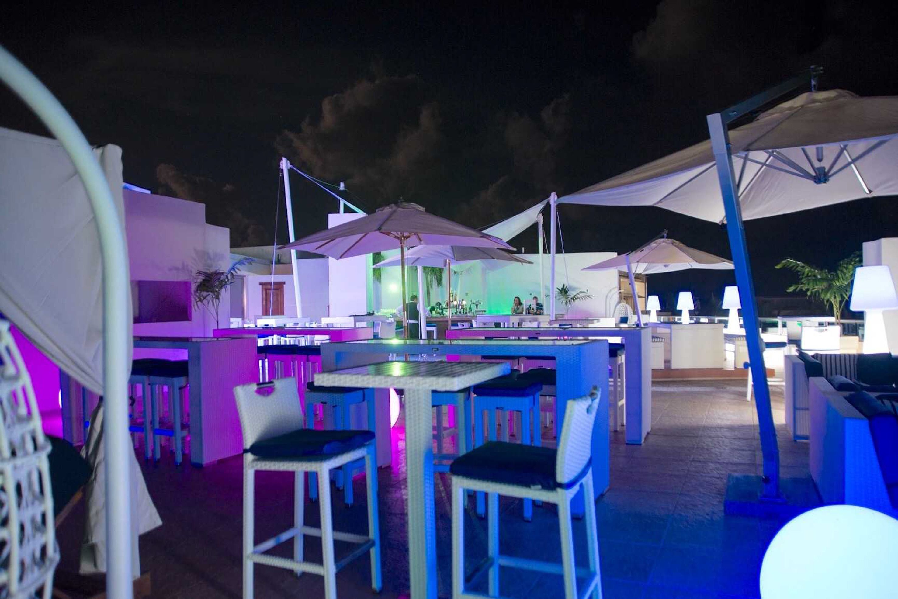 An outdoor bar area with tables and chairs, umbrellas, and a poolside setting at night.