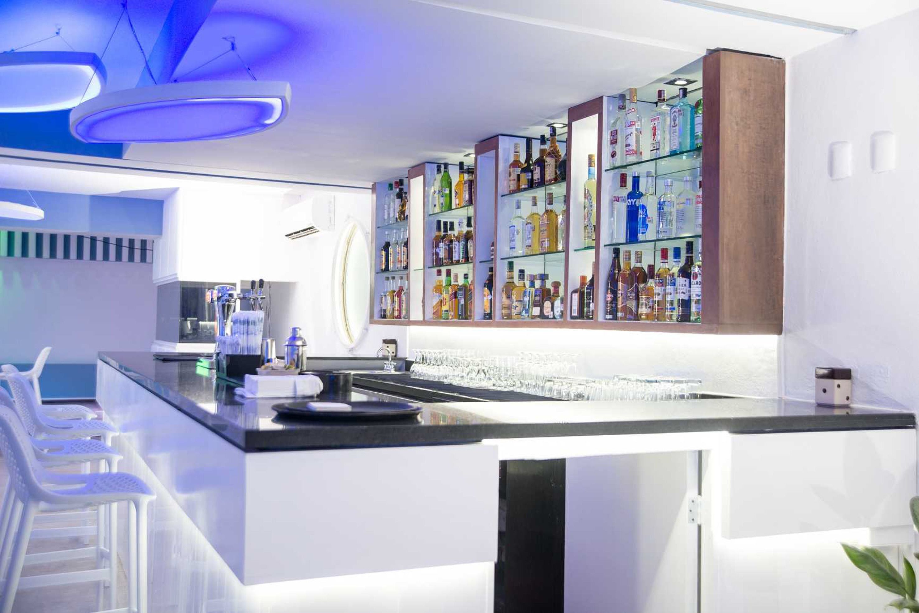 A modern bar area with a long counter, multiple shelves of liquor bottles, and white bar stools.