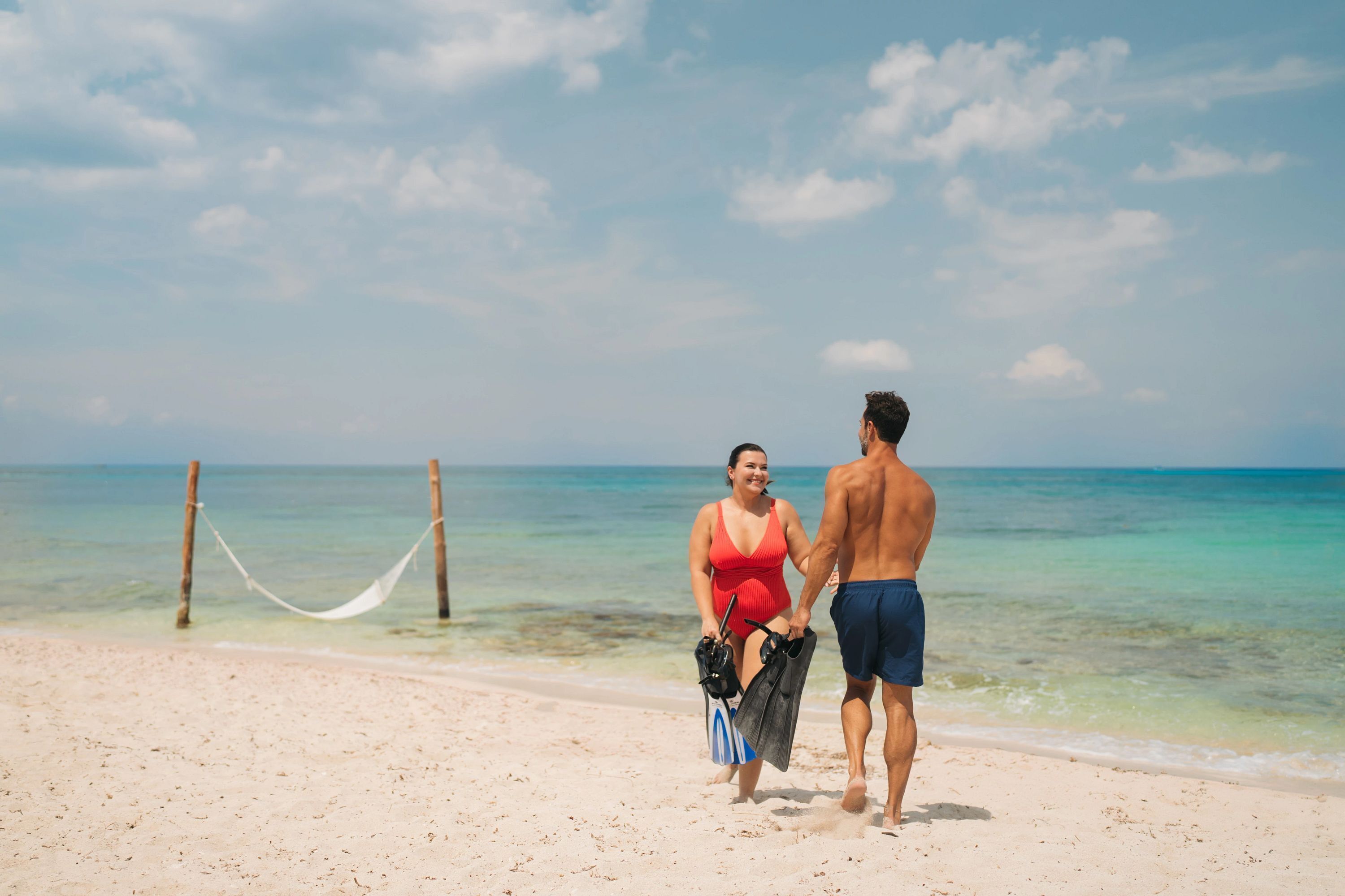 Un couple se promène sur une plage de sable, avec un hamac et une eau bleu clair en arrière-plan.