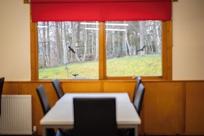 A room with a table and chairs, a window with a view of a forest, and a red blind.