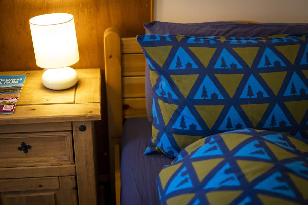 A wooden bedside table with a lamp, a book, and a patterned pillow on a bed.