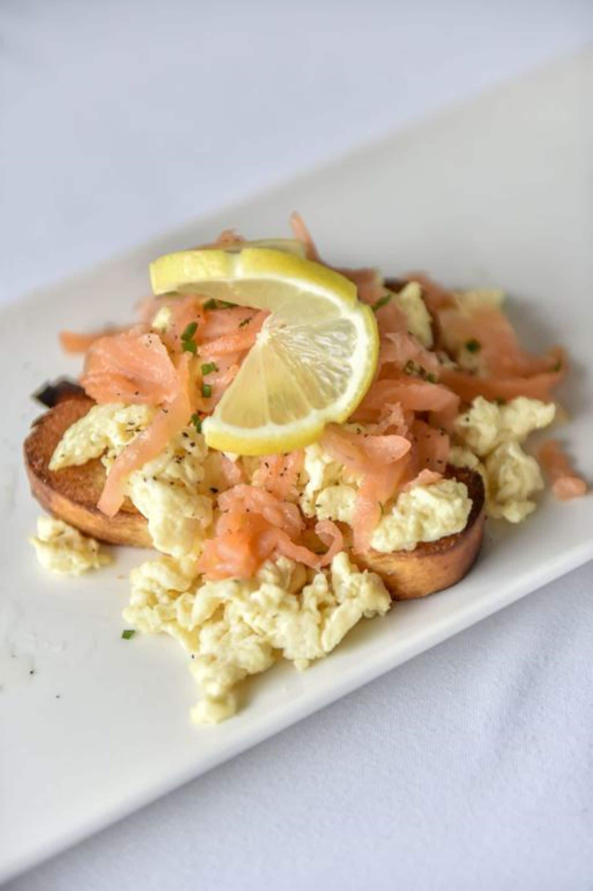 A dish with scrambled eggs, smoked salmon, and a lemon slice on a white plate.